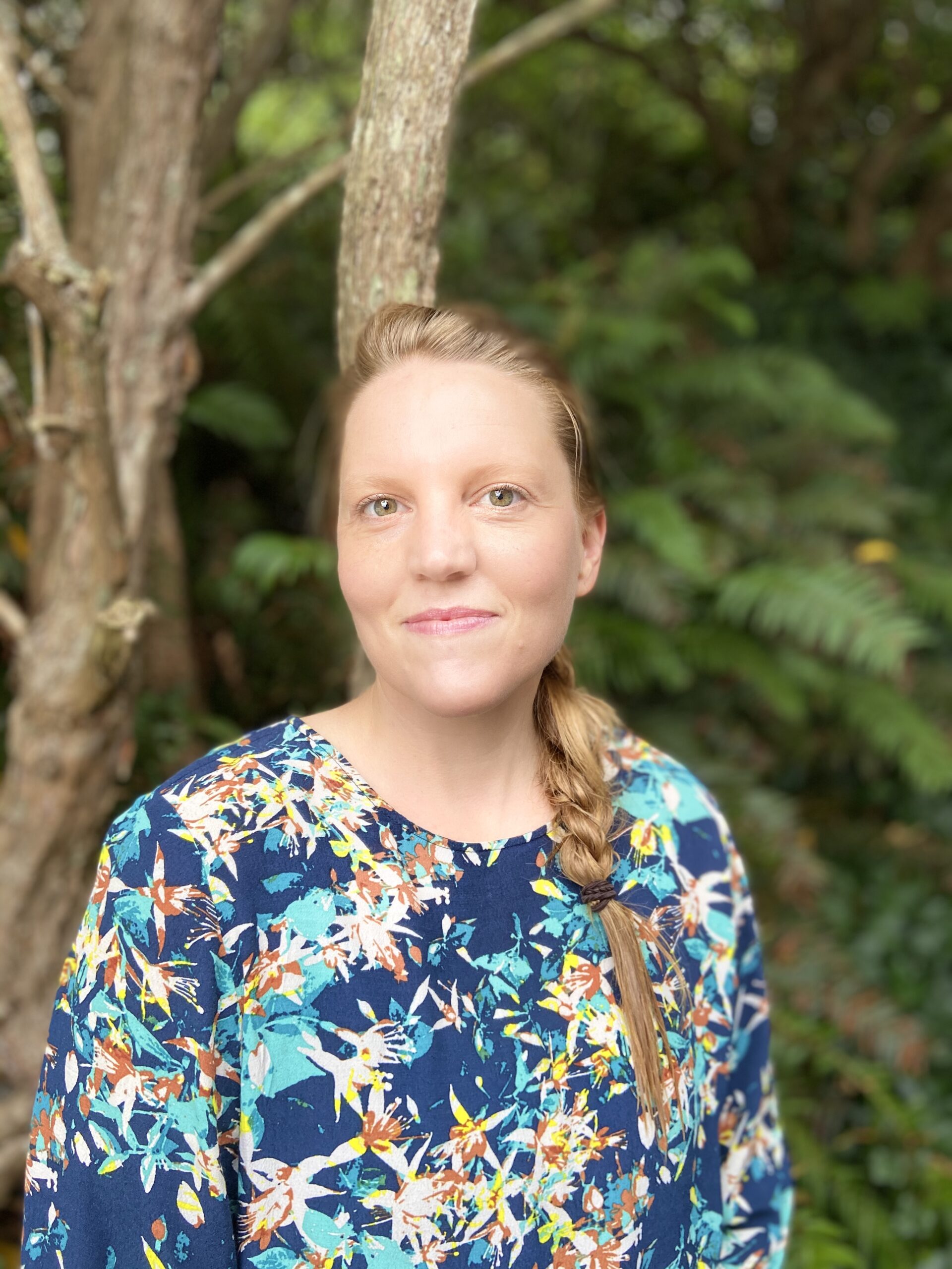 a light skinned woman with long red hair in a braid and a flowery shirt with a blue background stands in front of a background of ferns and trees while smiling gently